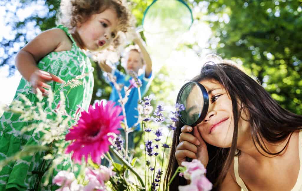 Three children explore a garden on a sunny day, surrounded by colourful flowers. One girl closely examines a purple bloom with a magnifying glass, while another child in a green dress looks on with curiosity. A third child in the background holds up a butterfly net, immersed in the joyful activity of outdoor discovery.