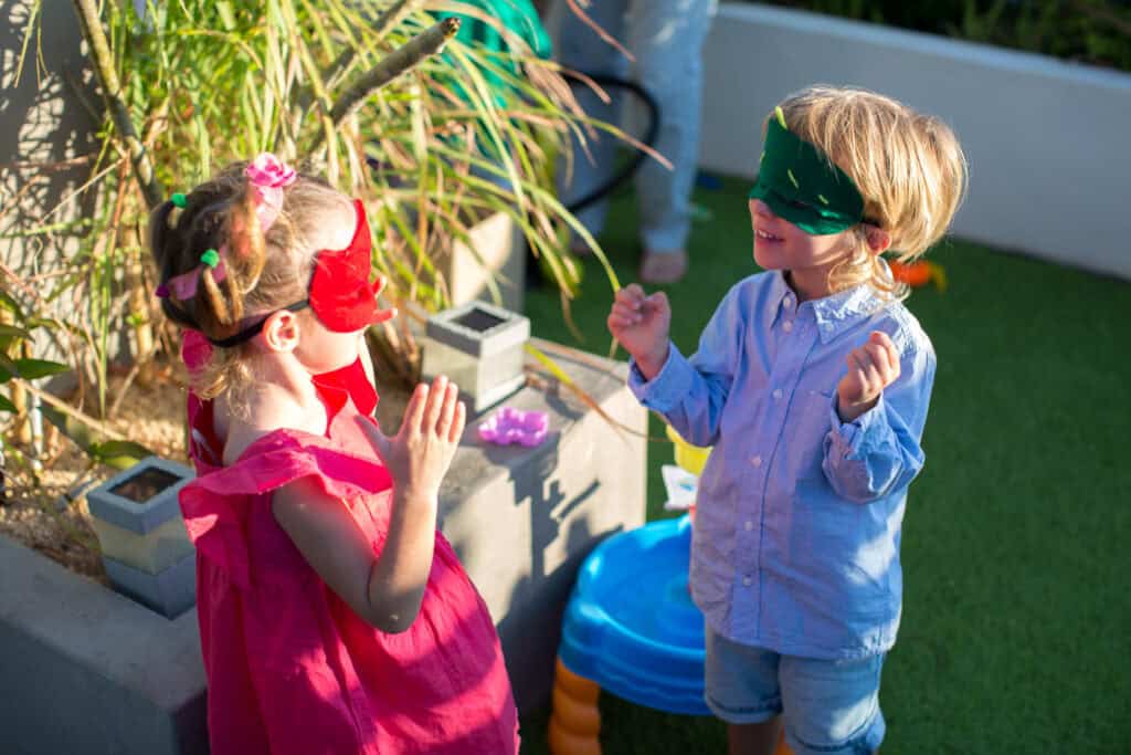 A girl and boy wearing colourful blindfolds laugh and interact during a party game in a sunny outdoor setting with playful decorations around them.