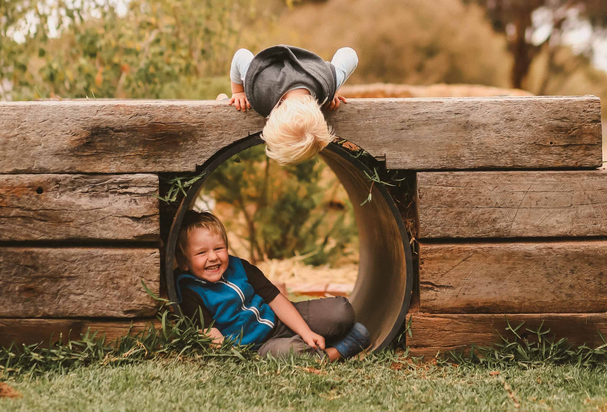 Two young children laugh and play around a wooden play tunnel—one child lies inside the tunnel smiling, while the other peers over the top upside down.