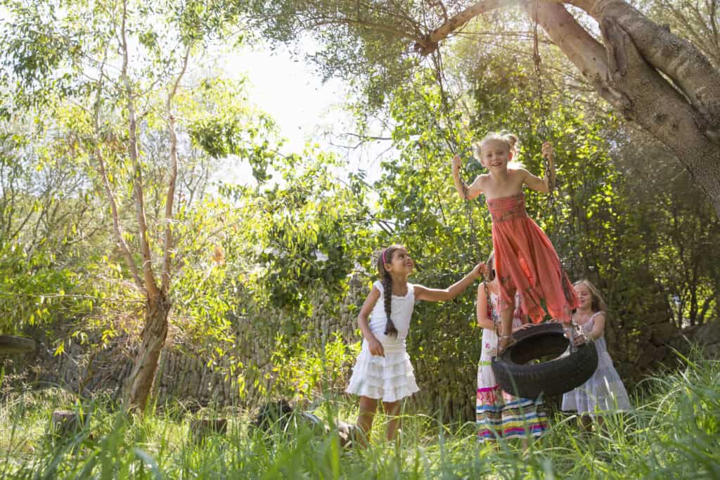Four girls are enjoying a sunny day in a lush, green outdoor space, with one child standing on a tire swing while the others help and cheer her on.