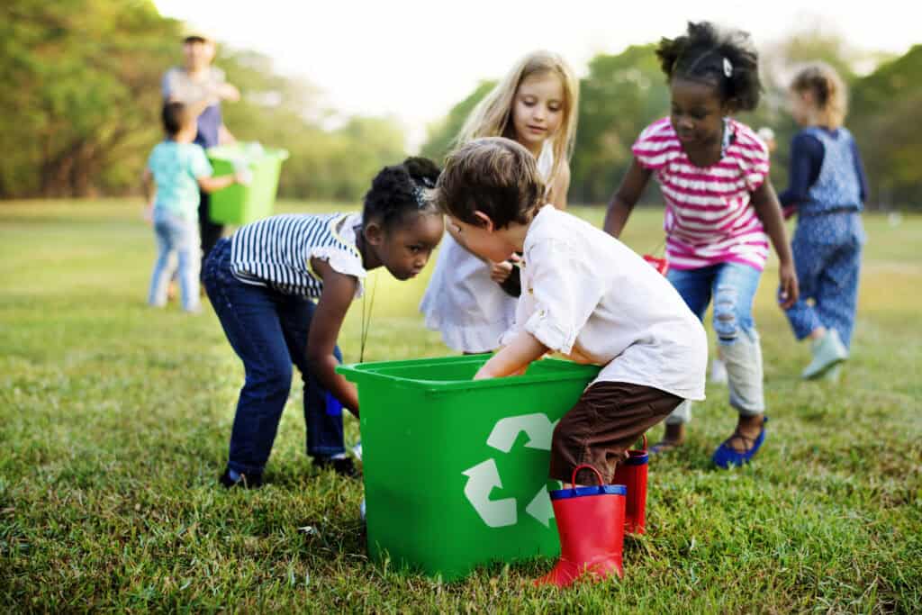 A group of young children collaborate to place items into a large green recycling bin on a grassy field, demonstrating teamwork and environmental care.