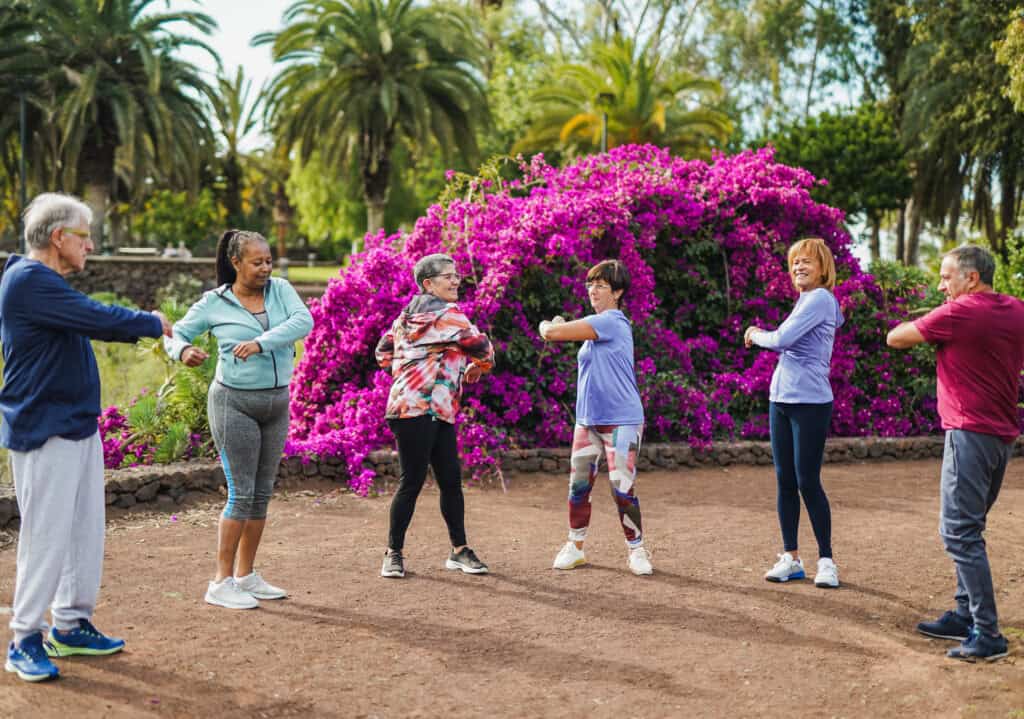 A diverse group of senior adults standing in a circle, engaging in stretching exercises in a park surrounded by vibrant pink flowers and palm trees.