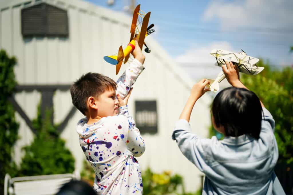 Two children enthusiastically hold up colourful toy airplanes, pretending to fly them in the garden on a sunny day.