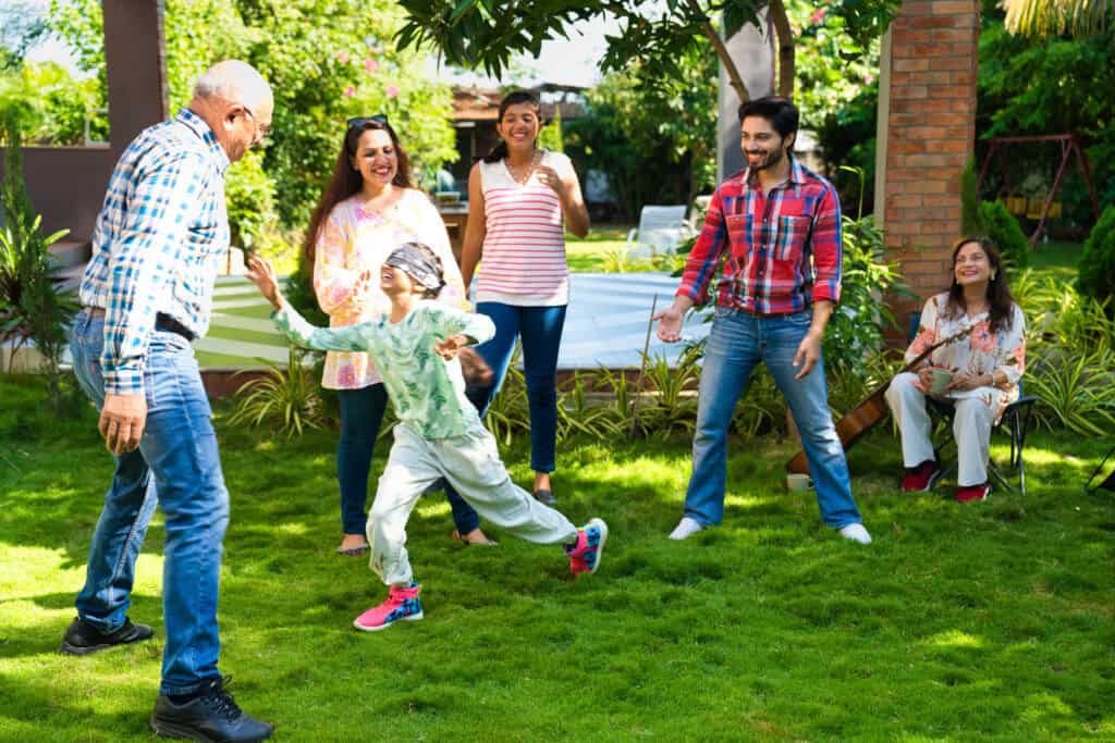 A joyful Indian family of six playing a blindfolded tag game in a lush green backyard, with grandparents, parents, and children all actively engaged.