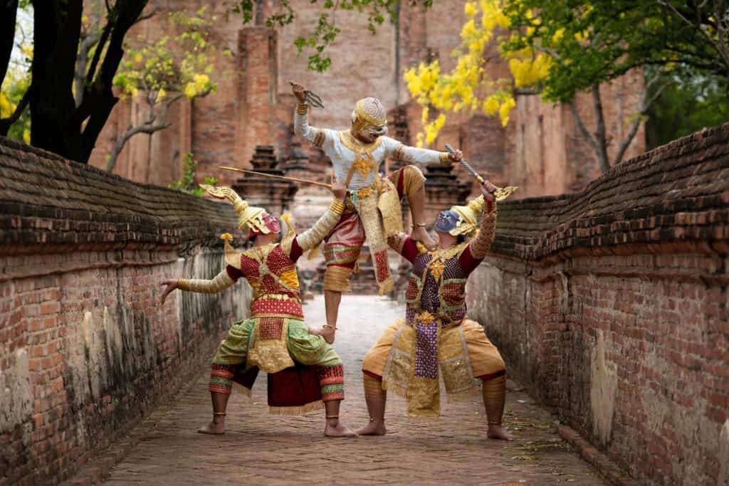 Three performers in ornate traditional Thai costumes and painted masks enact a dramatic pose during a classical Khon dance on a brick walkway surrounded by ancient temple walls.