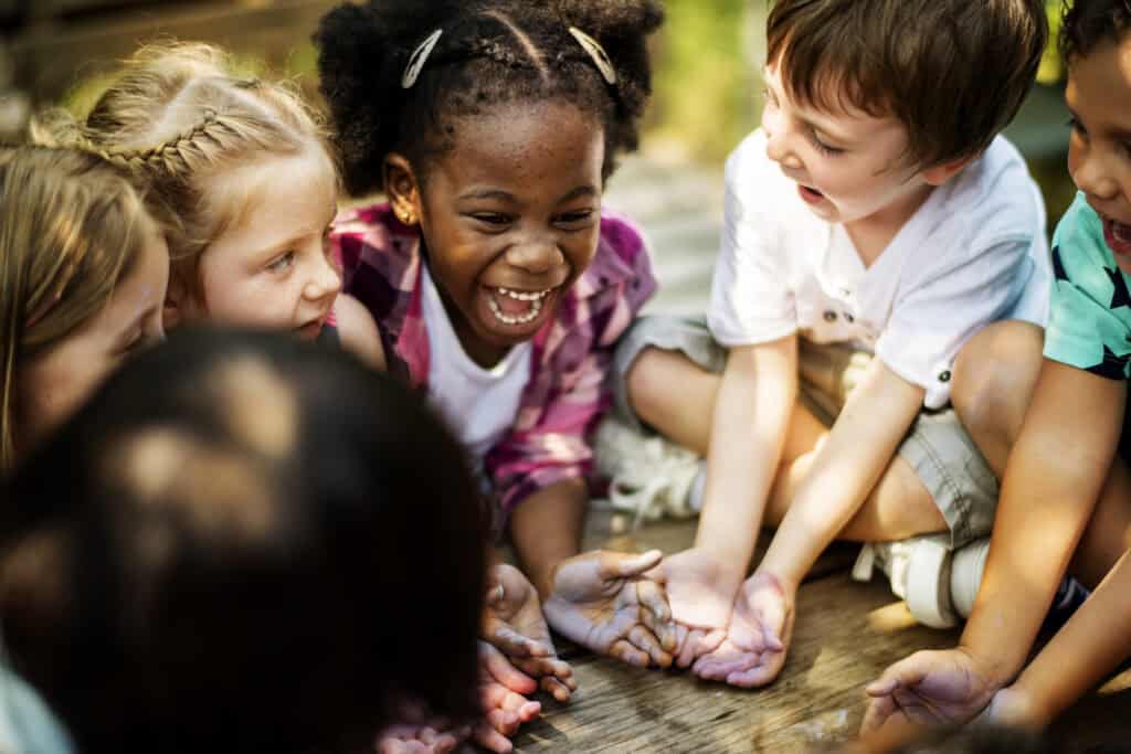 A joyful group of children sit in a circle, laughing and holding out their hands during a playful group activity.