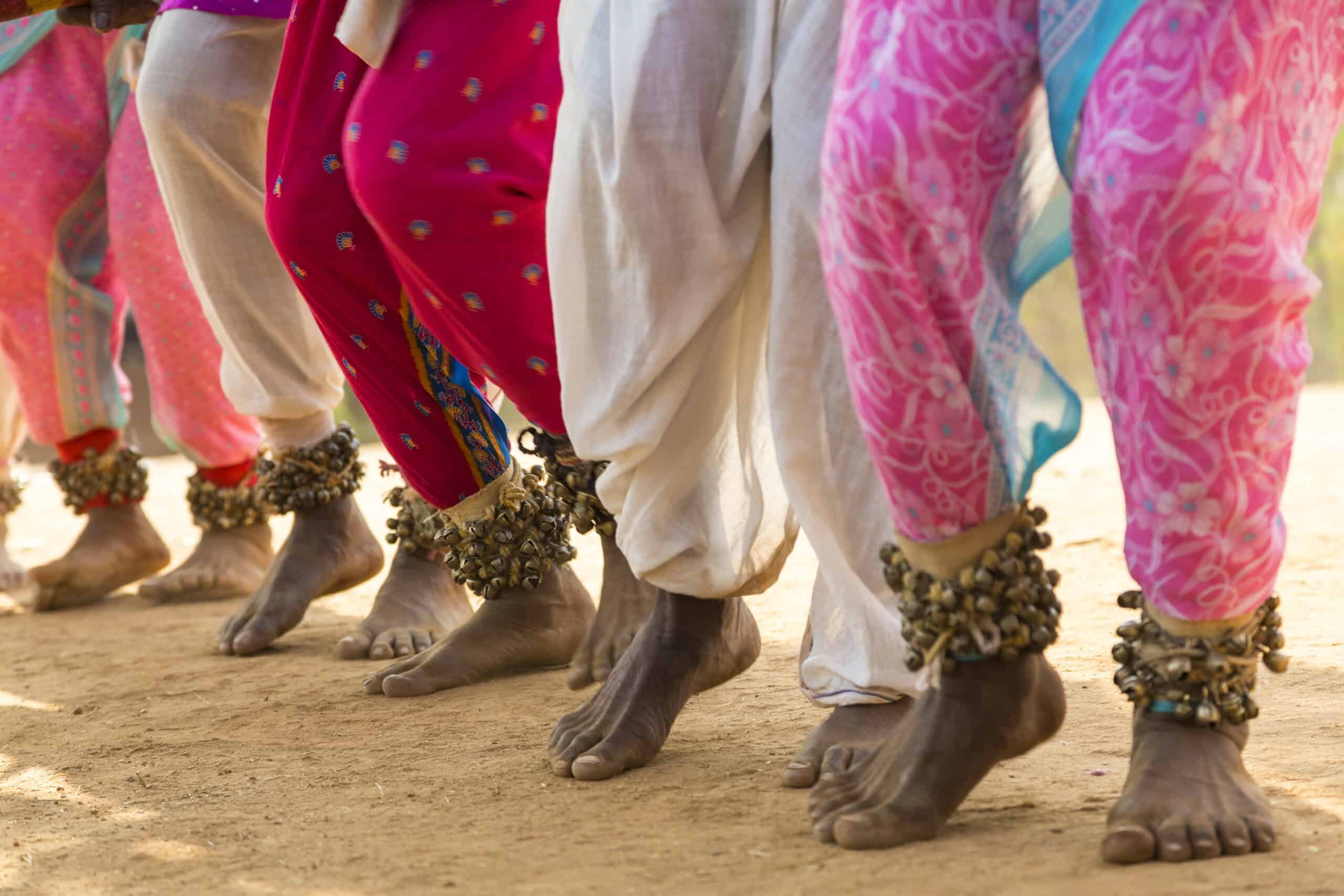 A close-up view of bare feet adorned with ankle bells, dancing in rhythm on a sandy surface, wearing colourful traditional trousers in shades of pink, red, and white.