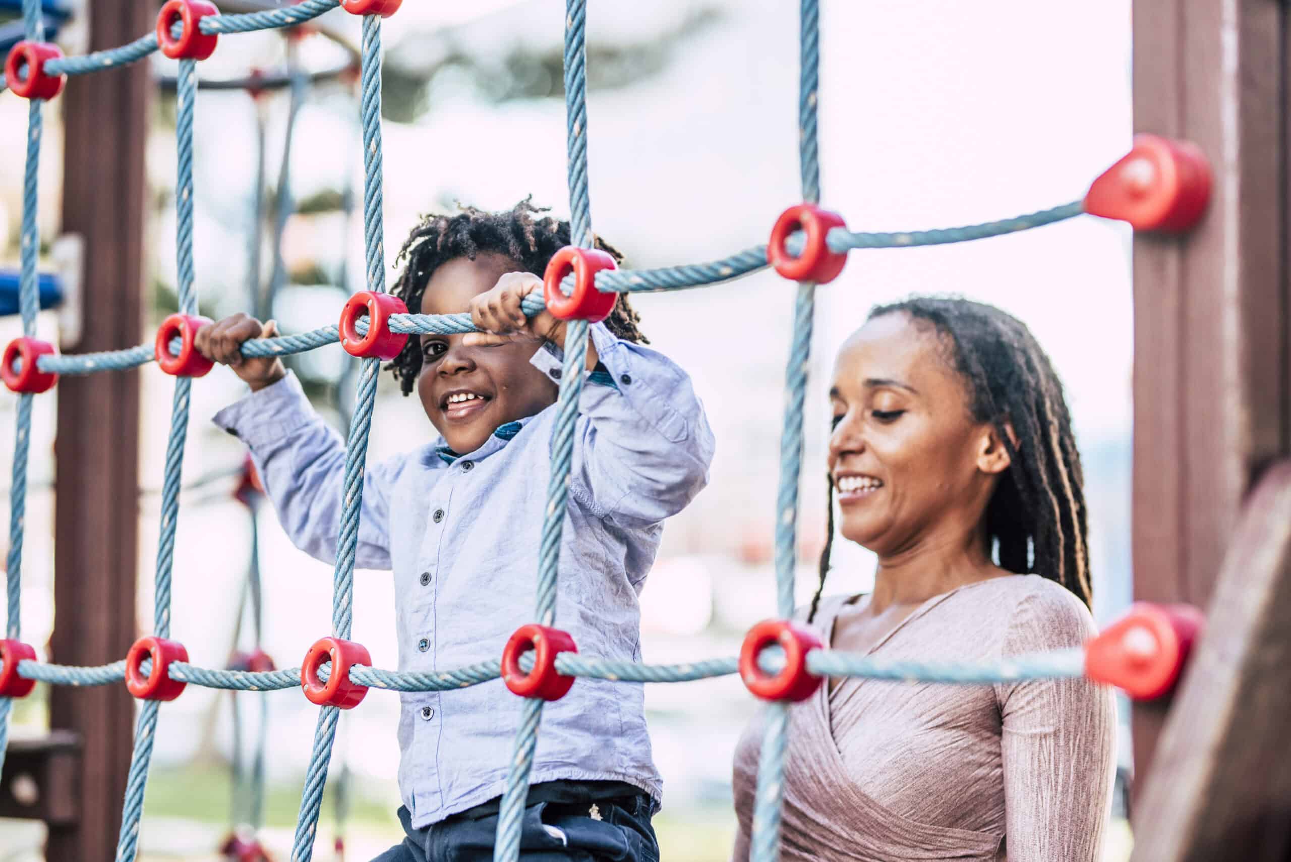 Smiling child climbing a rope structure with supportive mother beside him at a playground.