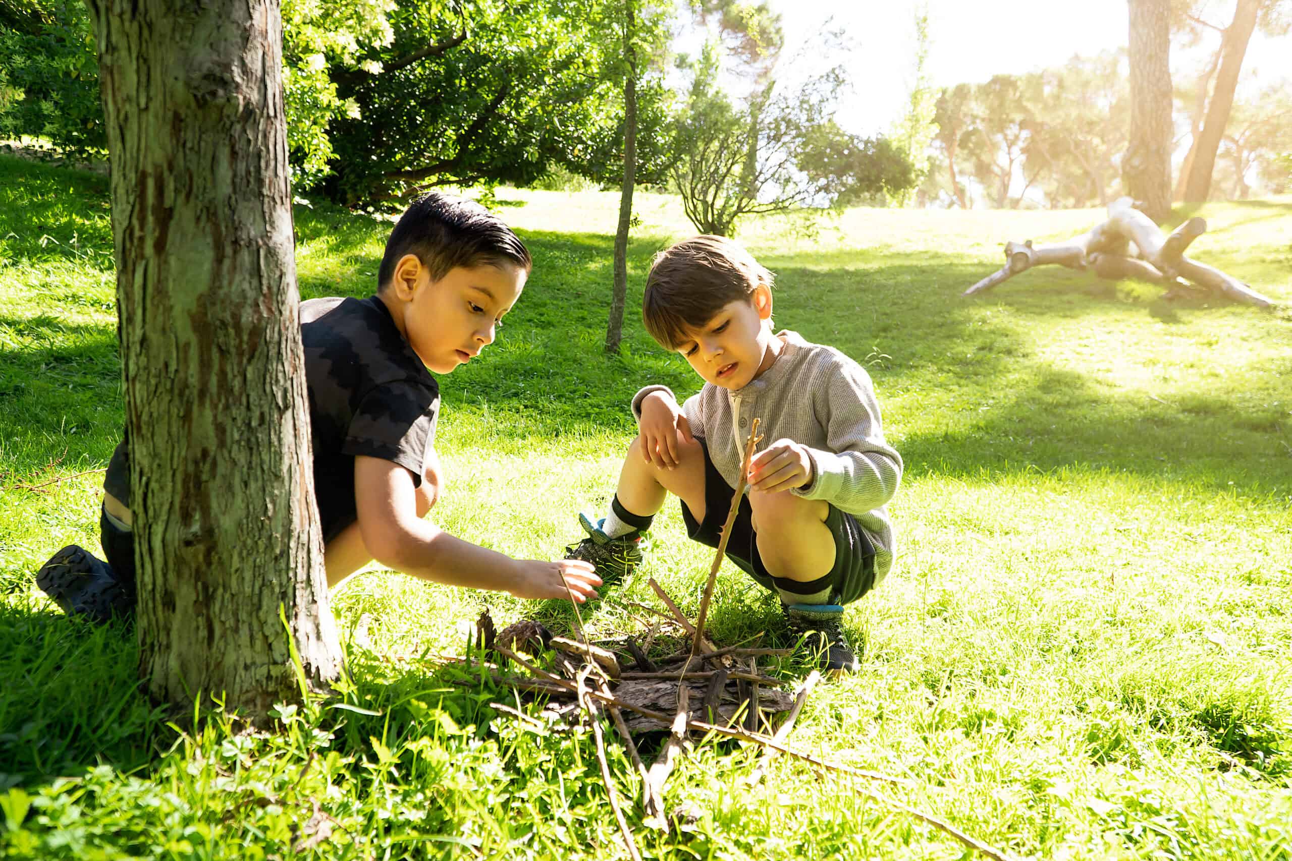 Two boys kneel on a sunlit patch of grass, carefully arranging sticks and bark to create a small structure in a natural play area.