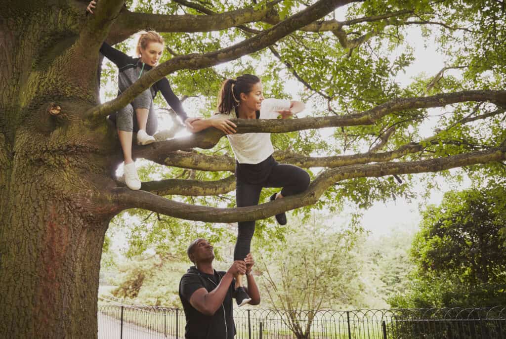 Two young women climbing a large tree with thick branches, one being assisted from below by a man in a supportive gesture in a green park setting.