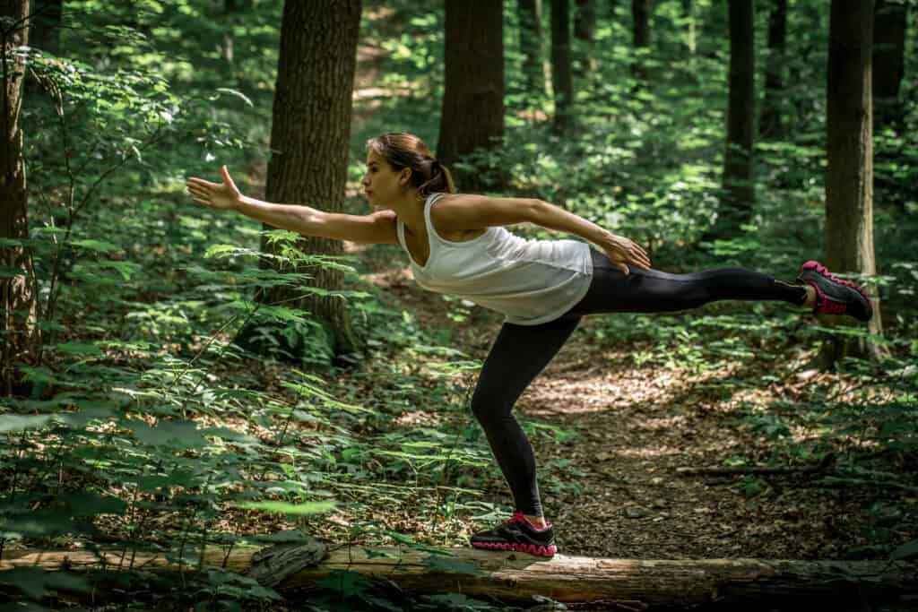 A woman balancing on one leg on a log in a dense green forest, extending one arm forward in a focused stretch during a mindful movement exercise.