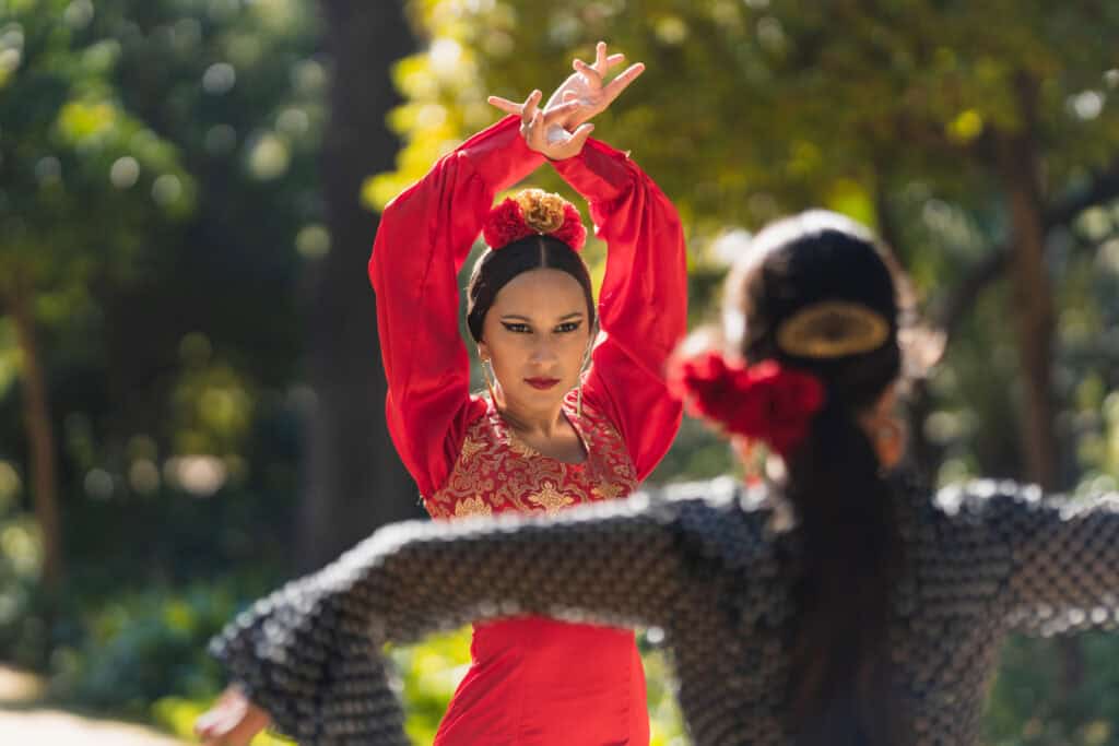 A woman in a red flamenco dress with flowers in her hair dances intensely, facing another dancer in traditional attire, with sunlight filtering through trees in the background.