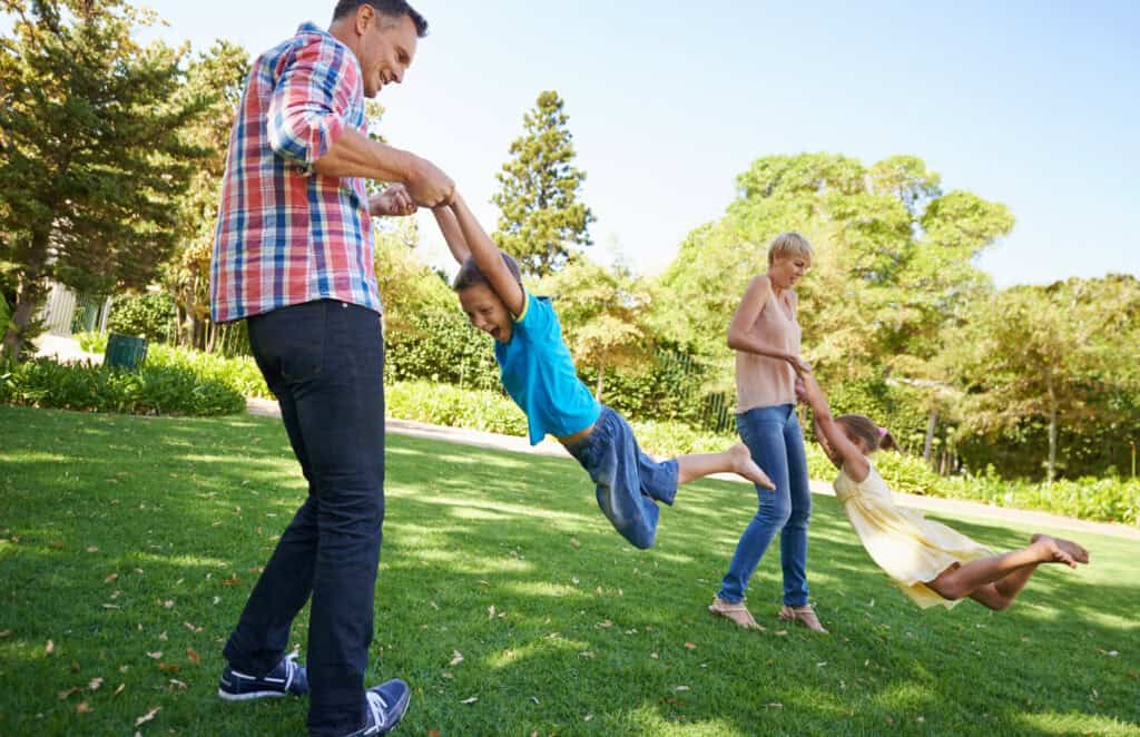 Two parents swing their laughing children by the arms in a park, enjoying energetic and playful outdoor family time.