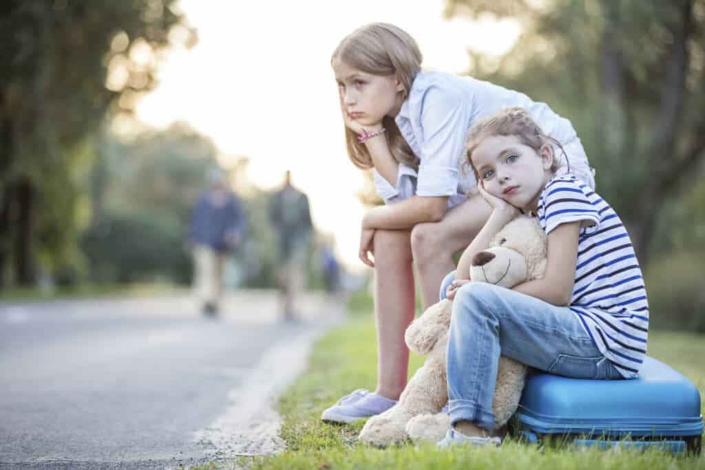 Two girls wait by the roadside at dusk: the older, in a light blue shirt and shorts, crouches pensively with her chin on her hand, while the younger sits on a small blue suitcase hugging a plush teddy bear, both looking tired as blurred figures walk in the distance.