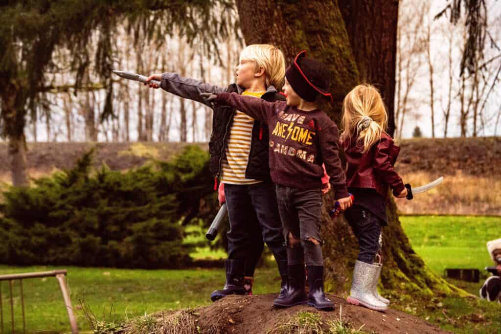 Three young children dressed as pirates pointing and playing together on a grassy hill under tall trees.