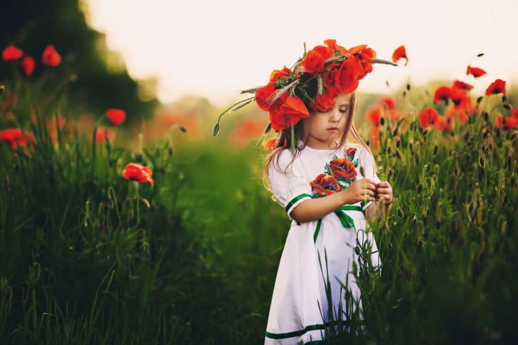 Young girl wearing a white dress with embroidered flowers and a crown of red poppies, standing in a lush green field surrounded by blooming poppies.