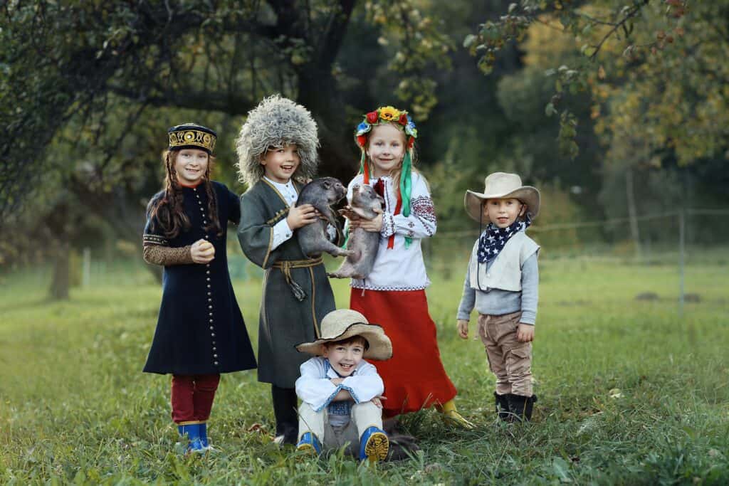 Group of diverse children dressed in traditional and folk costumes from different cultures, smiling and holding animals in a green field.