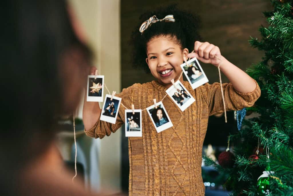 Smiling girl holding a string of instant photo prints in front of a decorated Christmas tree, showing off captured family moments.