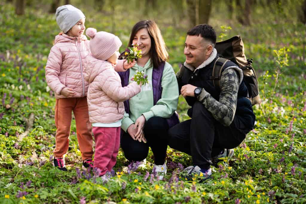 A family with two young girls in pink jackets pausing during a hike in a colorful, flower-filled forest—one child offering flowers to her smiling mother.