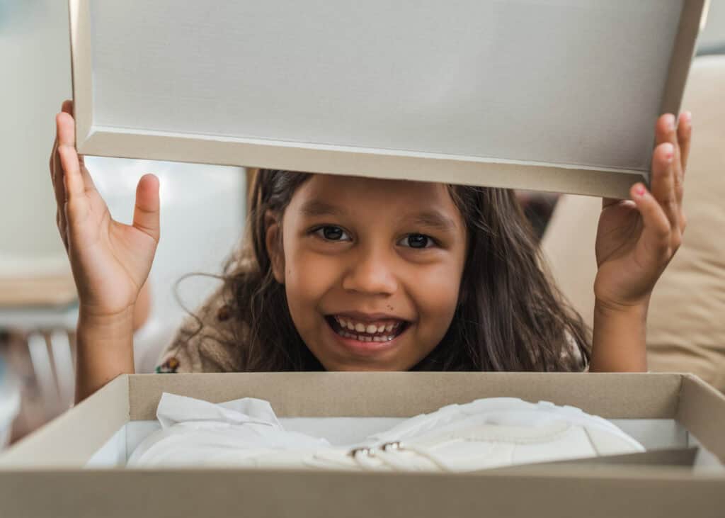 Smiling young girl lifting the lid of a cardboard box to reveal new white shoes inside, expressing excitement and joy.