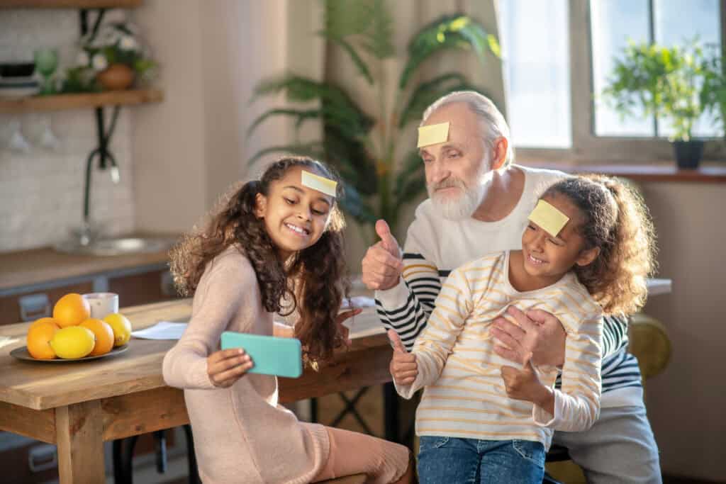 Grandfather and two young girls sitting at a kitchen table, smiling and playing a memory game with sticky notes on their foreheads while taking a selfie.