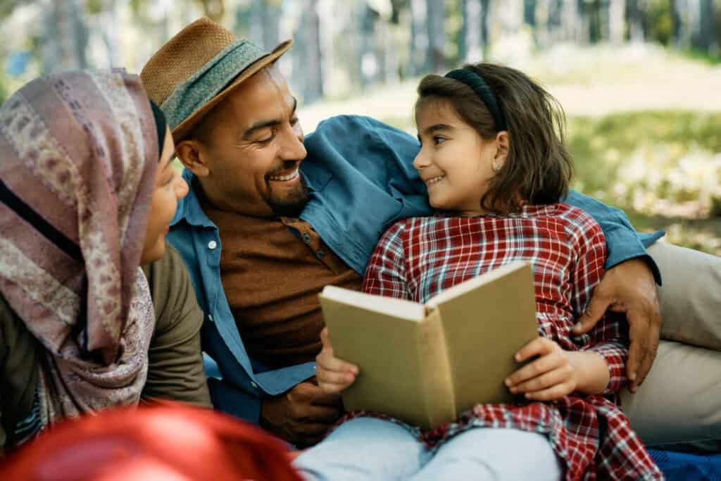 Family enjoying story time outdoors as a father and mother read a book with their smiling daughter in a natural, wooded environment.