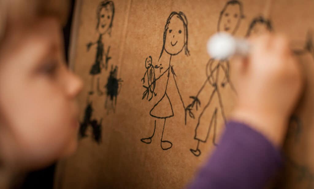 Child drawing a family portrait on cardboard using a black marker, focusing on simple stick figures and a doll-like character.