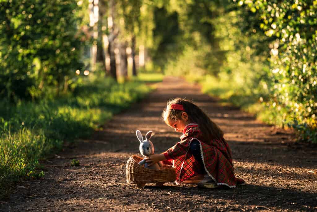 Little girl in a red plaid dress playing on a forest path, gently placing a toy bunny into a wicker cradle.