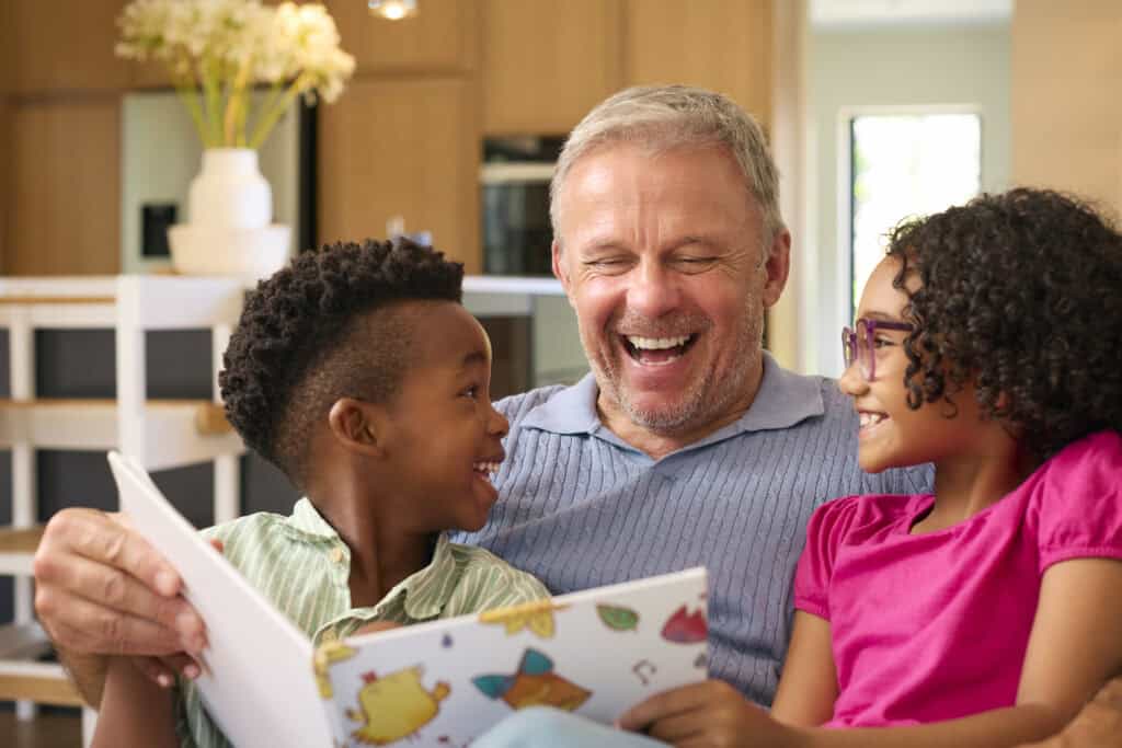 Grandfather joyfully reading a children’s book with a young boy and girl, all laughing together in a warm indoor setting.