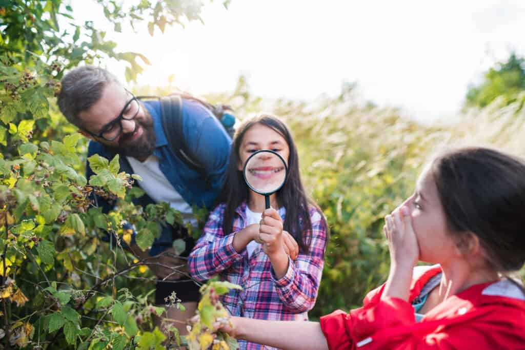 Smiling girl holding a magnifying glass over her mouth, creating a fun distorted smile, while a man and another child laugh beside her during a nature exploration.