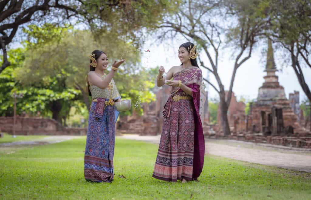 Two women dressed in traditional Thai clothing joyfully participating in a water-splashing ceremony in a historic park setting.