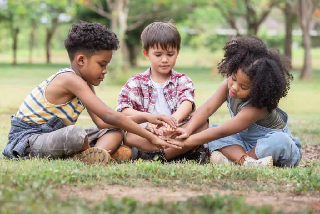 Three children sitting in a circle on grass, bonding through a hands-on nature activity.