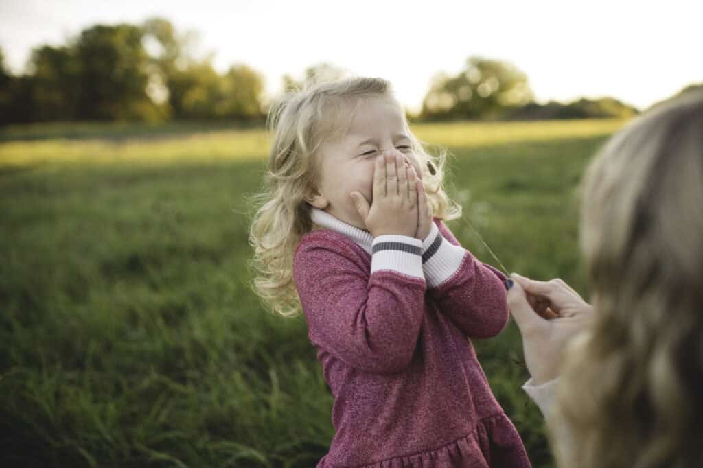 Blonde toddler girl giggling as an adult tickles her with a grass stem in a meadow.