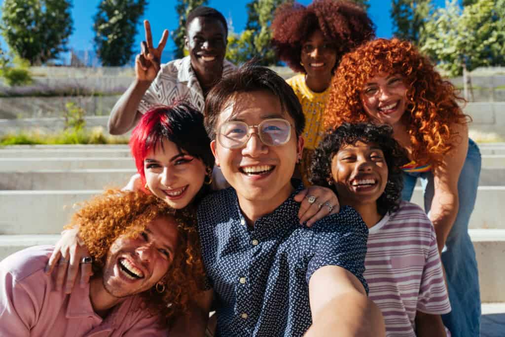 A joyful group of diverse young adults taking a cheerful selfie together on outdoor steps under a bright blue sky.