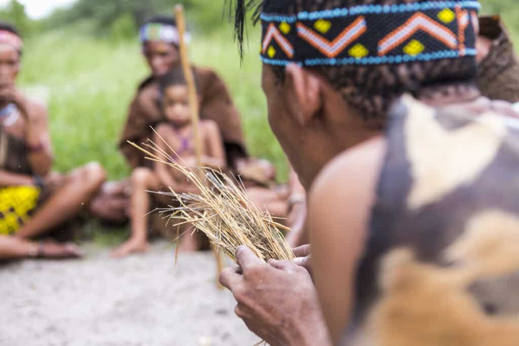 A member of the San people creating fire using dry kindling while a group, including a child, observes attentively in a natural outdoor setting.
