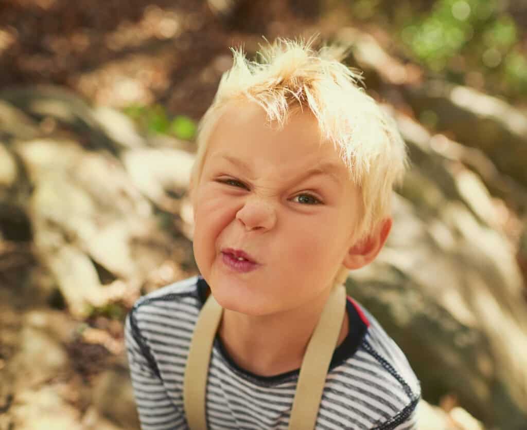 Blonde boy with tousled hair making a funny face outdoors.