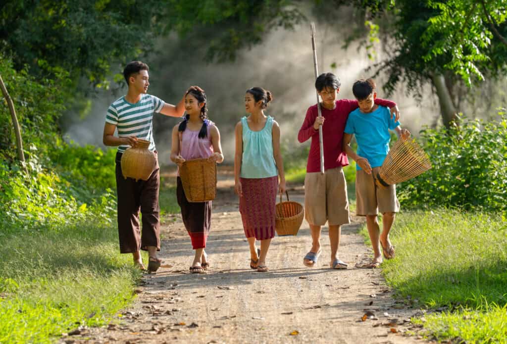 Five teenagers walking along a dirt path in a forest, carrying traditional fishing baskets and tools, enjoying time together.