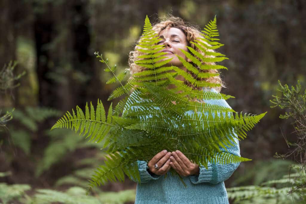 A woman in a turquoise sweater gently holds large green fern fronds close to her face, eyes closed, surrounded by forest greenery.