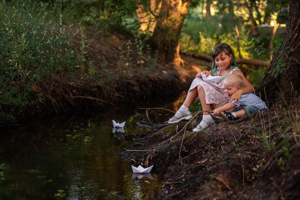 An older sister and her little brother sit by the water’s edge, launching paper boats into the calm stream.