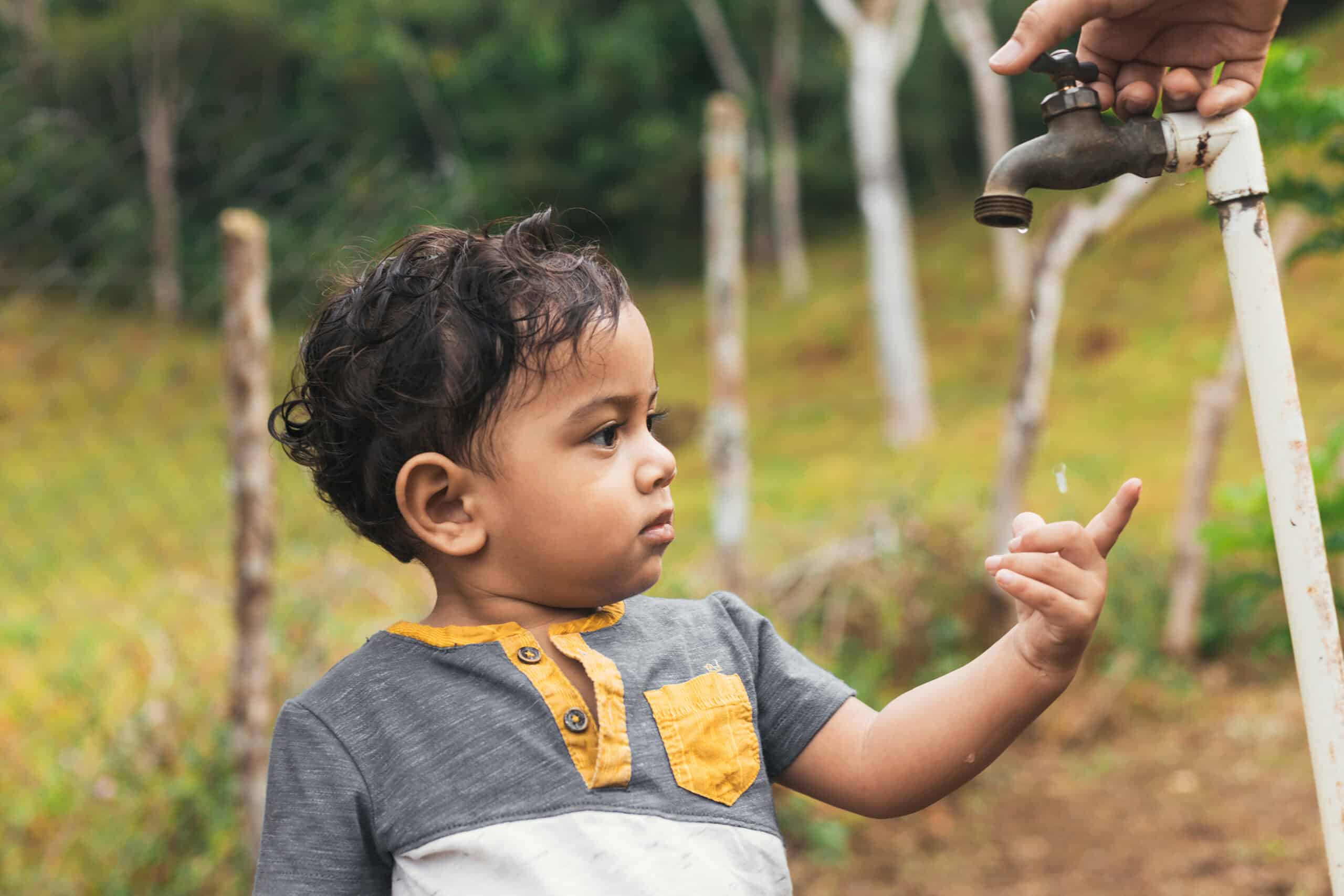 A young boy watches droplets fall from an outdoor tap, reaching out his finger to catch them.