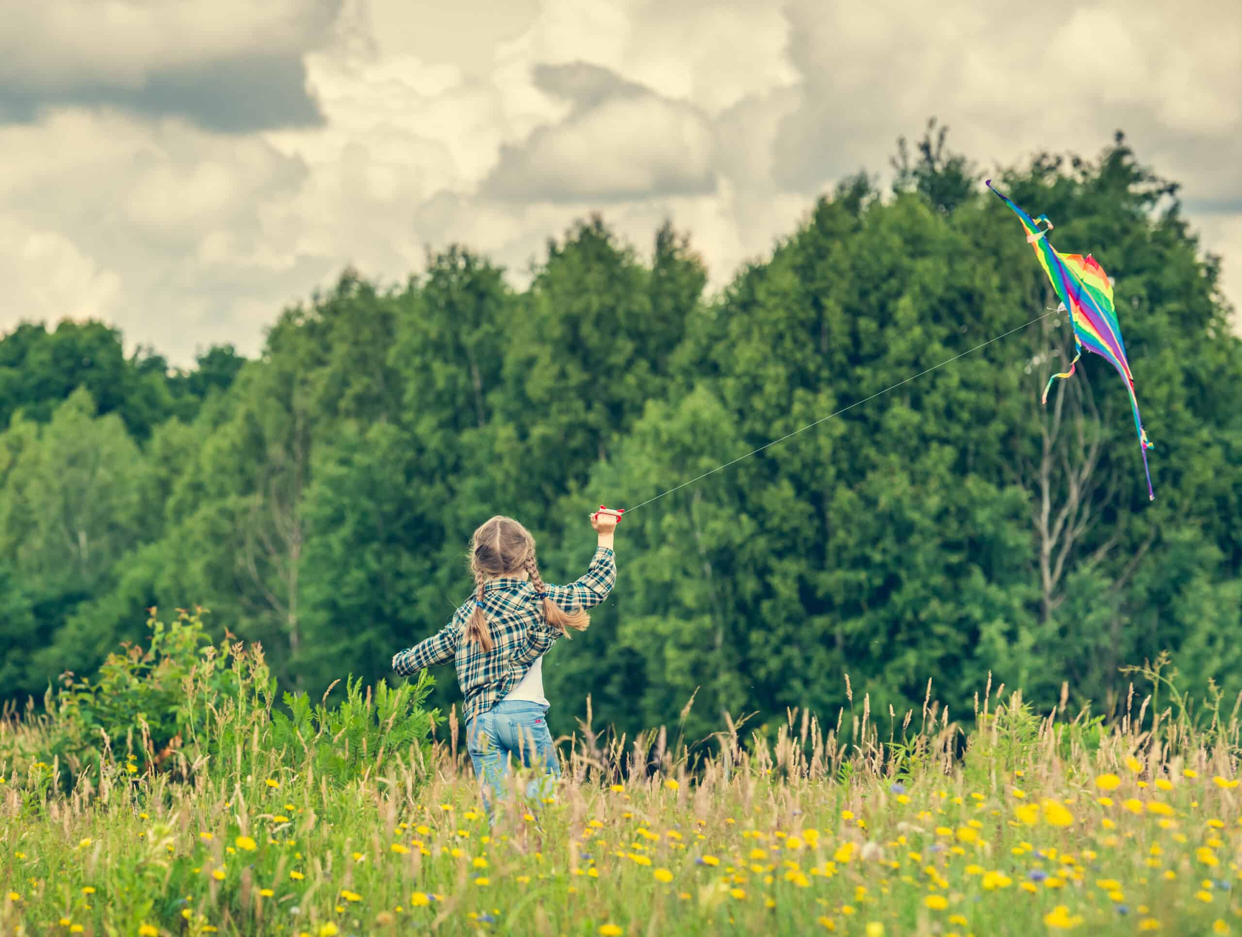 A girl with braided hair runs through a wildflower field, flying a rainbow kite against a backdrop of trees and clouds.
