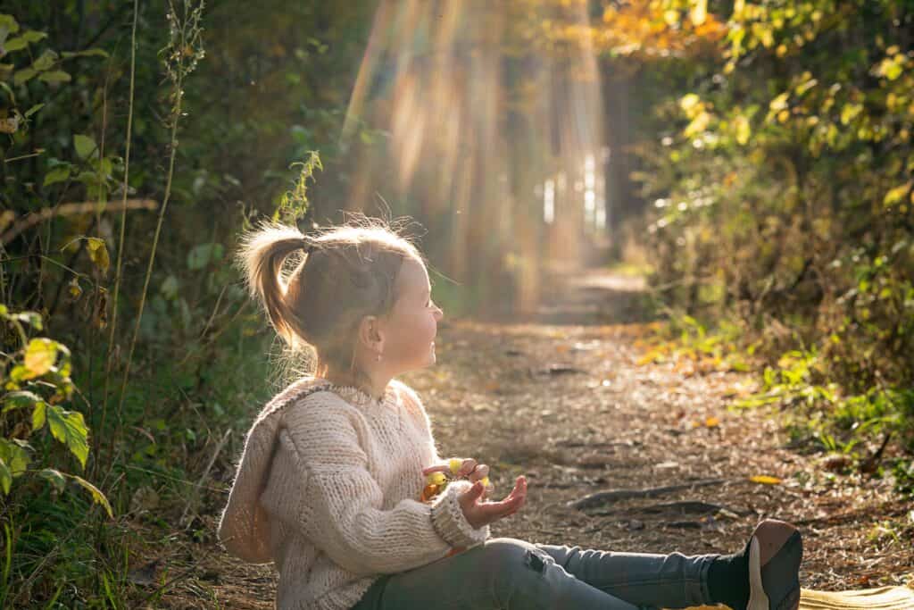 A small girl with a ponytail and a cozy sweater sits on a wooded trail, smiling as sunlight streams through the trees.
