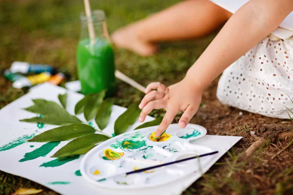 Close-up of a child’s hand dipping fingers into green and yellow paint beside leaves and art supplies spread out on the grass.