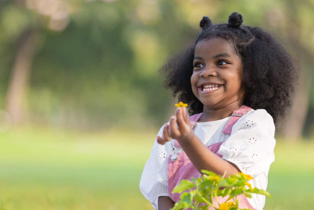 A joyful child in pink overalls and a white shirt smiles brightly while holding a small yellow flower outdoors.