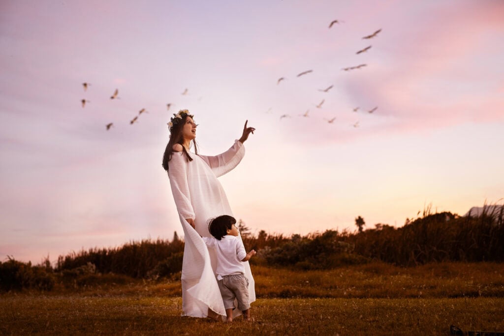 A woman wearing a flower crown and white dress points toward a flock of birds in the pink evening sky as a young child stands beside her, holding her dress.