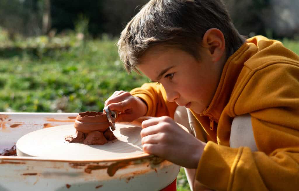 A focused boy in an orange jumper carefully molds a clay pot on a spinning wheel set up outdoors in natural light.