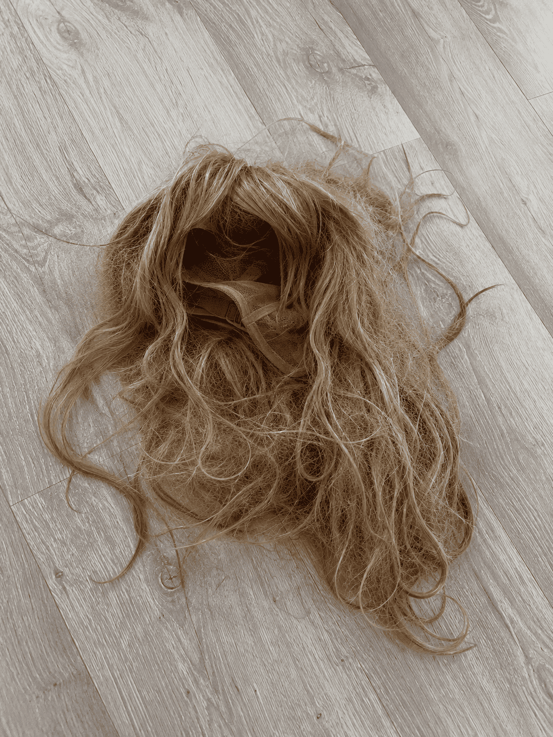 A frizzy, messy light brown synthetic wig discarded on a grey wood floor