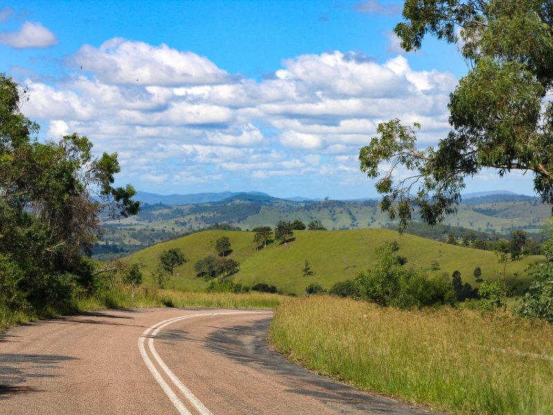 A scenic rural road winding through lush green hills with trees on both sides, under a bright blue sky with scattered clouds, showcasing the peaceful countryside of New South Wales.