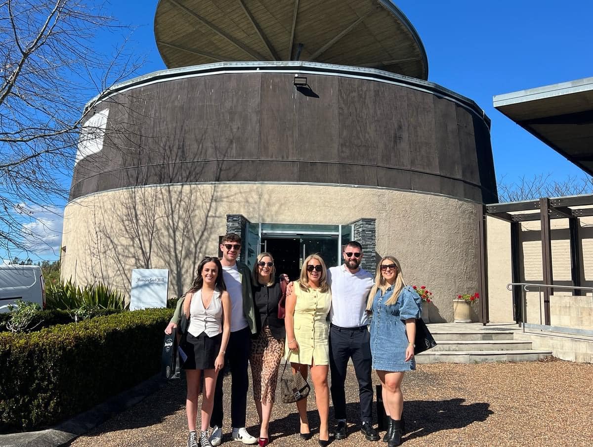 Explore a group of six friends enjoying a wine tasting tour at a Hunter Valley winery, with clear blue skies and scenic vineyard views in the background.