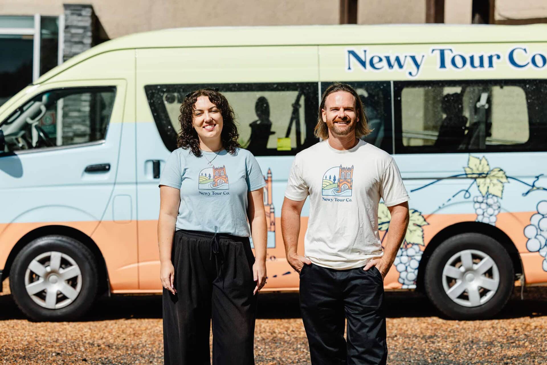 Vibrant Newy Tour Co. branded minibus with two smiling staff members in casual attire standing in front, showcasing local tours and wine tasting experiences in Newcastle.